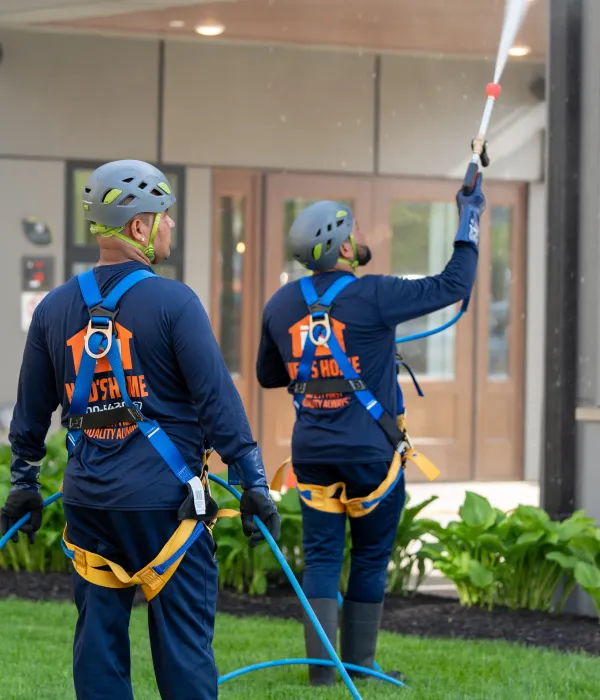 Two professional technicians in safety gear pressure-wash warehouse windows during industrial facility cleaning service