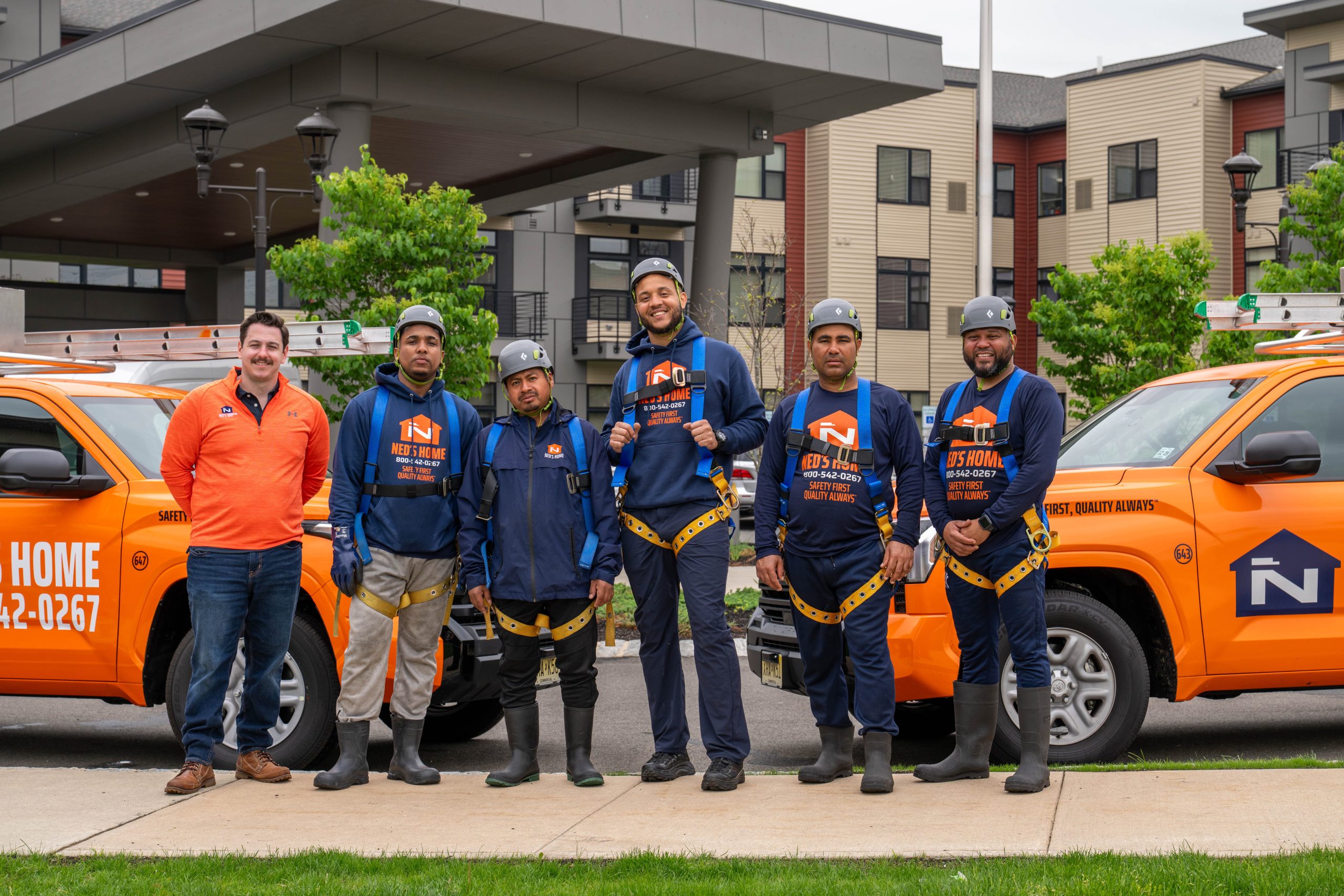 Ned's Home commercial services crew in uniforms and safety harnesses standing in front of service trucks outside a multi-unit residential property.