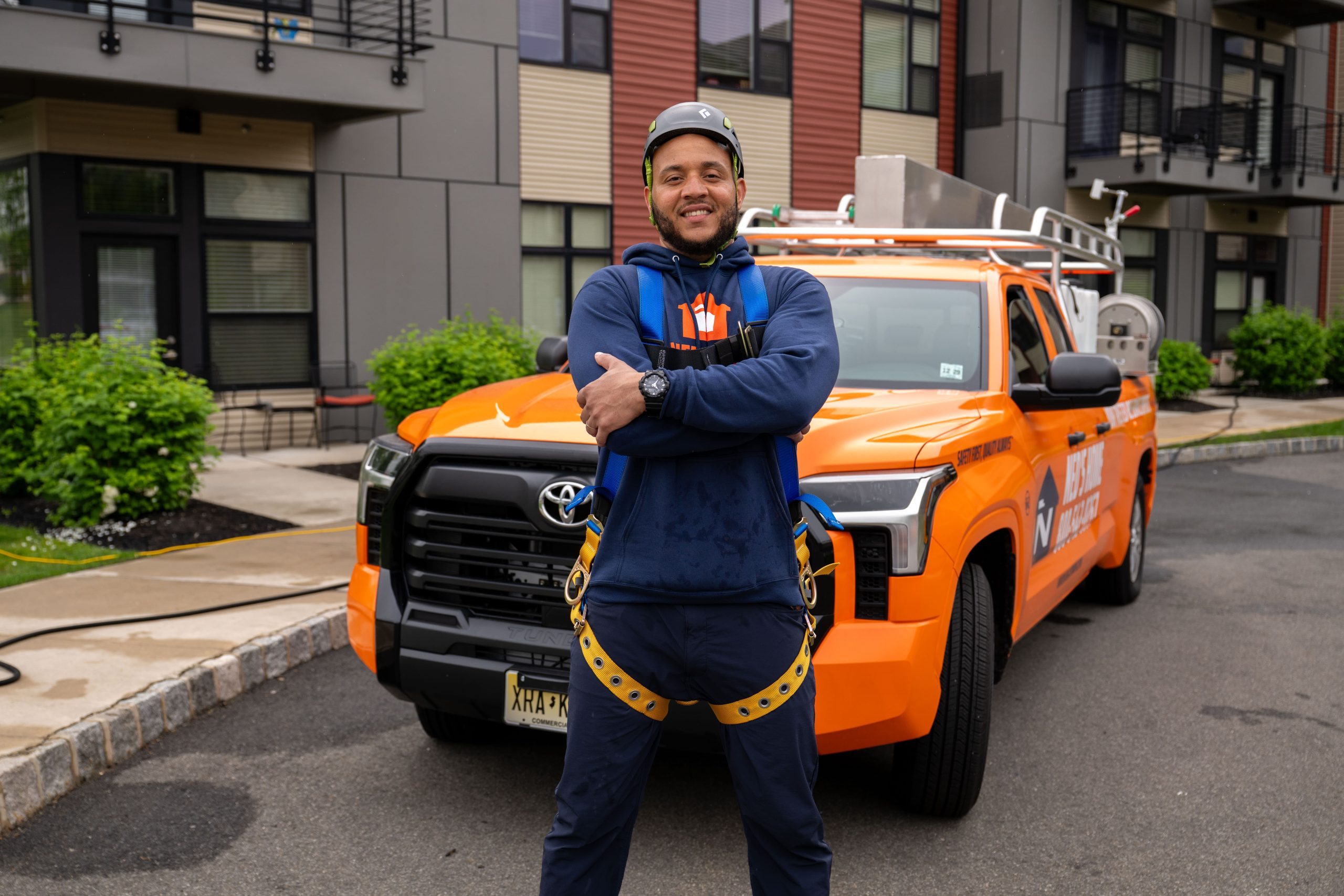 Ned's Home technician in safety harness and branded uniform standing in front of a service truck outside a commercial multi-unit residential property.